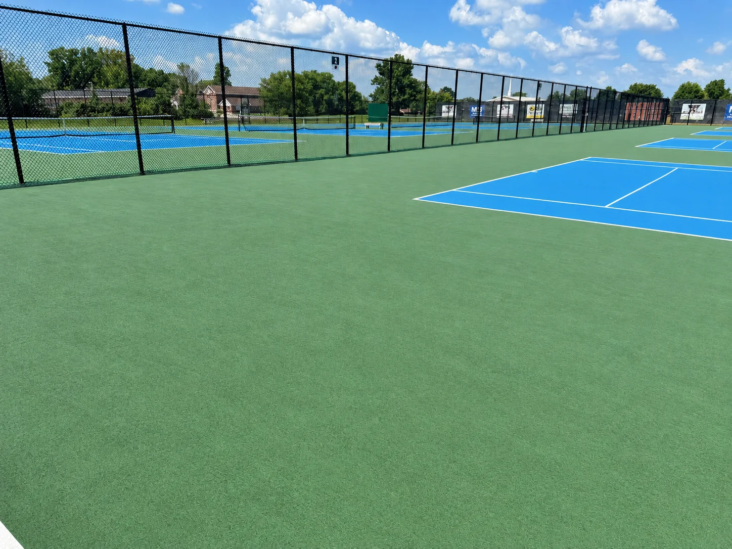 Cleaned green tennis court in the foreground with a blue court beyond and the black chain-link fence along the complex