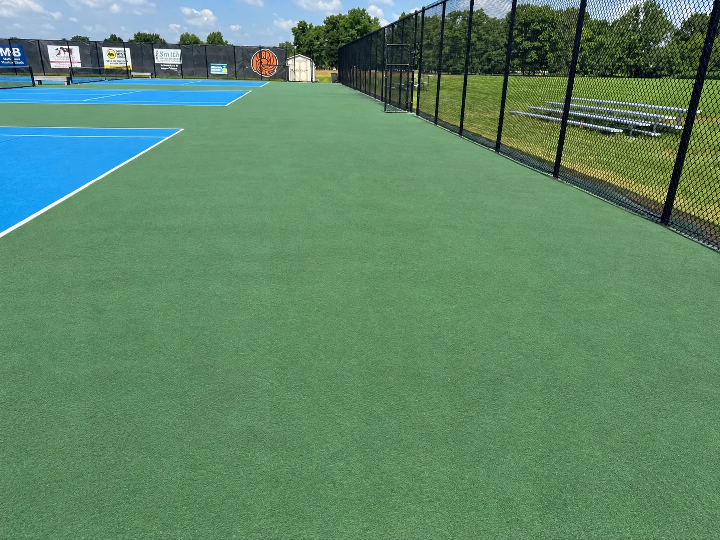 Long view down the cleaned tennis complex between the fence and the courts, with bleachers on the grass outside