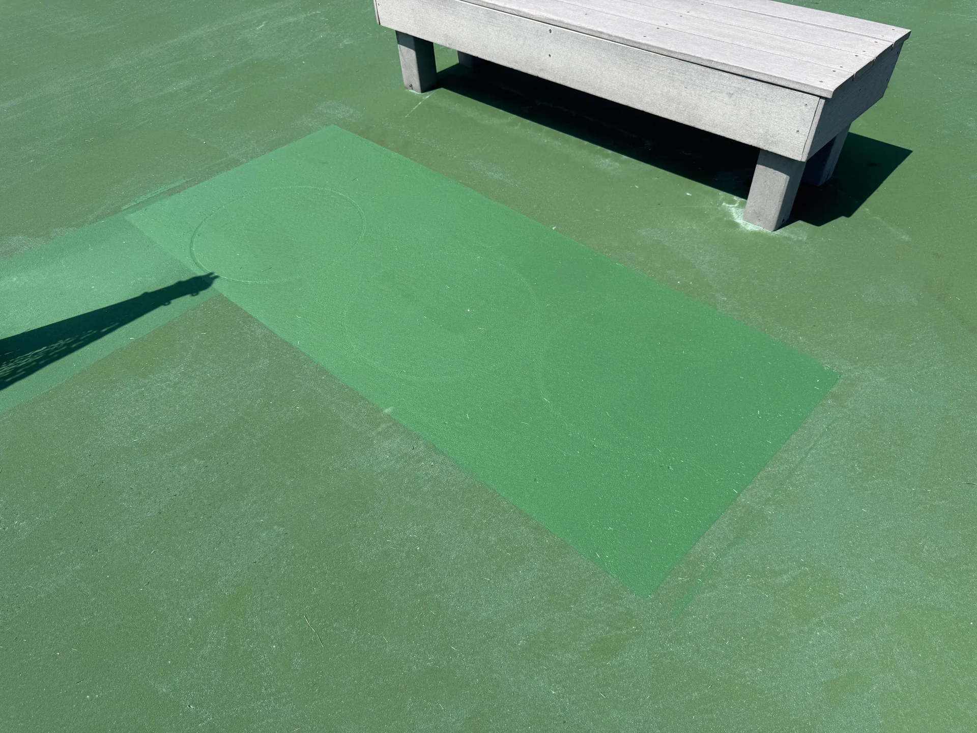 Bright rectangle of cleaned green court surface beside a bench, with faint circular marks from the rotary washer