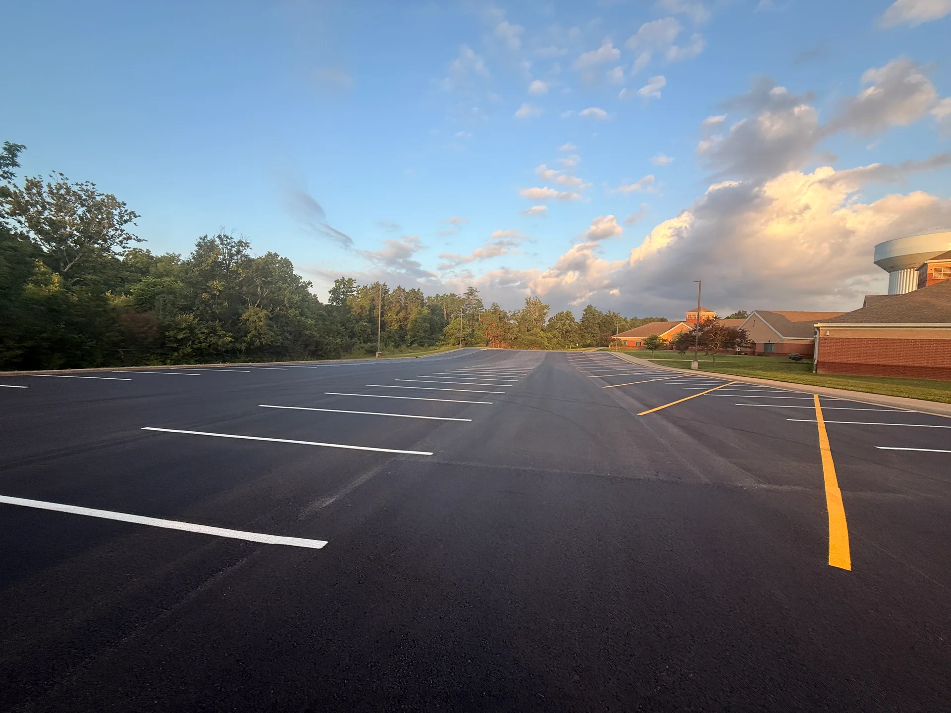 Freshly paved and striped school parking lot with yellow lane markings, a school building and water tower in the distance
